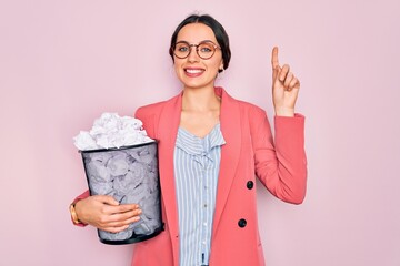 Wall Mural - Young beautiful business woman with blue eyes holding heap bin of crumpled papers surprised with an idea or question pointing finger with happy face, number one