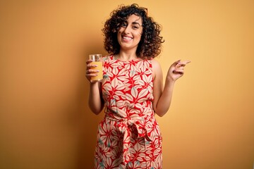 Poster - Young beautiful curly arab woman drinking glass of healthy orange juice over yellow background very happy pointing with hand and finger to the side