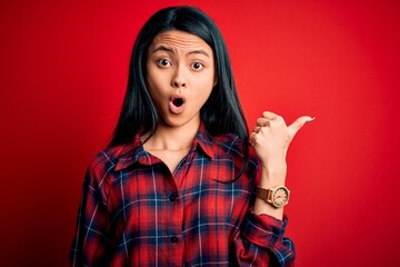 Wall Mural - Young beautiful chinese woman wearing casual shirt over isolated red background Surprised pointing with hand finger to the side, open mouth amazed expression.