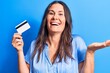 Young beautiful brunette woman holding credit card over isolated blue background celebrating achievement with happy smile and winner expression with raised hand