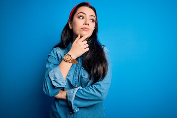 Wall Mural - Young brunette woman wearing casual denim shirt over blue isolated background with hand on chin thinking about question, pensive expression. Smiling with thoughtful face. Doubt concept.