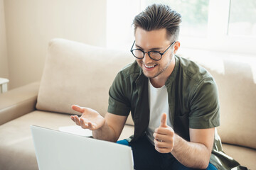 Bearded caucasian boy with eyeglasses having an online meeting at the laptop at home sitting on sofa