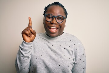 Sticker - Young african american plus size woman with braids wearing casual sweatshirt and glasses pointing finger up with successful idea. Exited and happy. Number one.