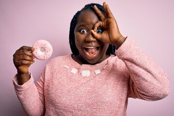 Sticker - Young african american plus size woman with braids eating doughnut over pink background with happy face smiling doing ok sign with hand on eye looking through fingers