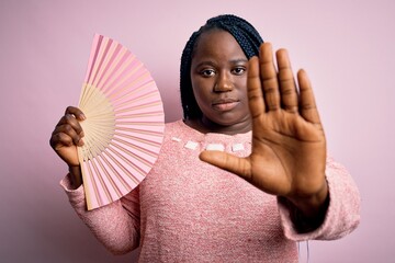 Poster - Young african american plus size woman with braids using hand fan over pink background with open hand doing stop sign with serious and confident expression, defense gesture