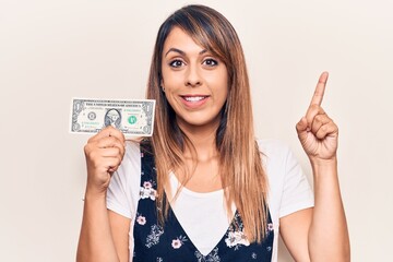 Young beautiful woman holding one dollar smiling with an idea or question pointing finger with happy face, number one