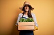© Krakenimages.com - Young beautiful brunette gardener woman wearing apron and hat holding box with plants with a happy face standing and smiling with a confident smile showing teeth