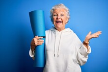 Senior Beautiful Sporty Woman Holding Mat For Yoga Standing Over Isolated Blue Background Very Happy And Excited, Winner Expression Celebrating Victory Screaming With Big Smile And Raised Hands