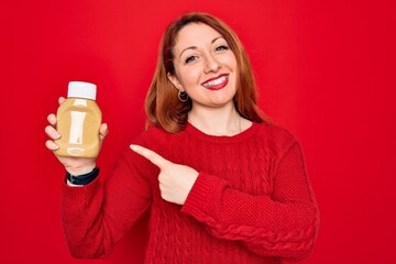 Canvas Print - Beautiful redhead woman holding bottle of mustard sauce condiment over red background very happy pointing with hand and finger