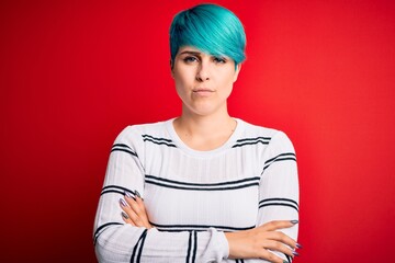 Young beautiful woman with blue fashion hair wearing striped sweater over red background skeptic and nervous, disapproving expression on face with crossed arms. Negative person.