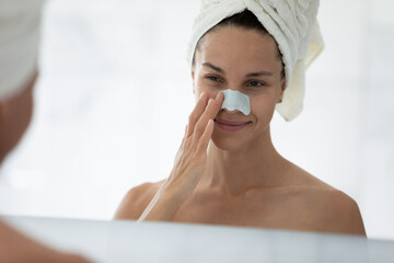 Wall Mural - Mirror reflection close up young woman applying anti blackhead patch, beautiful girl wearing white bath towel on head using nose strip mask, standing in bathroom after shower, skin care treatment