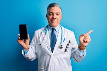 Poster - Middle age handsome grey-haired doctor man holding smartphone showing screen very happy pointing with hand and finger to the side