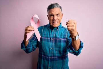 Wall Mural - Middle age handsome grey-haired man holding pink cancer ribbon over isolated background annoyed and frustrated shouting with anger, crazy and yelling with raised hand, anger concept