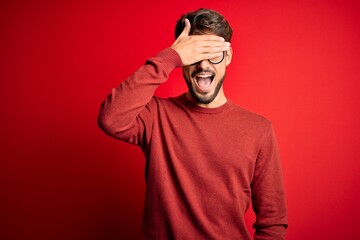 Poster - Young handsome man with beard wearing glasses and sweater standing over red background smiling and laughing with hand on face covering eyes for surprise. Blind concept.