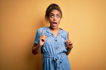 Poster - Young beautiful african american girl wearing denim dress standing over yellow background amazed and surprised looking up and pointing with fingers and raised arms.