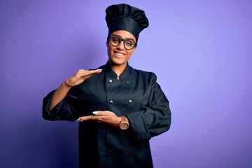 Poster - Young african american chef girl wearing cooker uniform and hat over purple background gesturing with hands showing big and large size sign, measure symbol. Smiling looking at the camera. Measuring