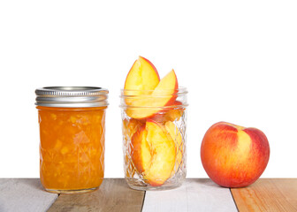 Fresh sliced peaches in an open mason jar with whole peach on table next to a full jar of home made canned peach jam on light colored wood table, isolated on white background.