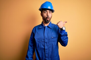 Poster - Mechanic man with beard wearing blue uniform and safety helmet over yellow background Surprised pointing with hand finger to the side, open mouth amazed expression.