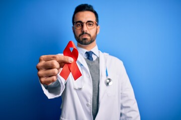 Poster - Young handsome doctor man with beard wearing stethoscope holding red hiv ribbon with a confident expression on smart face thinking serious