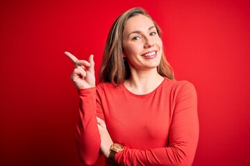 Wall Mural - Young beautiful blonde woman wearing casual t-shirt standing over isolated red background with a big smile on face, pointing with hand and finger to the side looking at the camera.