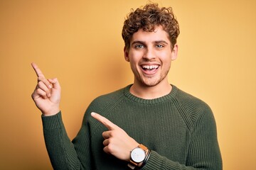 Young blond handsome man with curly hair wearing green sweater over yellow background smiling and looking at the camera pointing with two hands and fingers to the side.