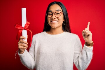 Poster - Young beautiful graduate asian woman holding university degree diploma over red background surprised with an idea or question pointing finger with happy face, number one