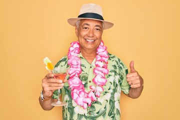 Middle age senior grey-haired man wearing summer hat and hawaiian lei drinking a cocktail happy with big smile doing ok sign, thumb up with fingers, excellent sign