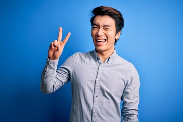 Wall Mural - Young handsome chinese man wearing casual shirt standing over isolated blue background smiling with happy face winking at the camera doing victory sign with fingers. Number two.