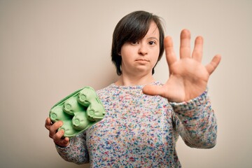 Poster - Young down syndrome woman holding cardboard egg cup from fresh healthy eggs with open hand doing stop sign with serious and confident expression, defense gesture