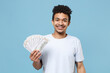 © ViDi Studio - Smiling young african american guy in casual white t-shirt isolated on pastel blue background studio portrait. People lifestyle concept. Mock up copy space. Hold fan of cash money in dollar banknotes.
