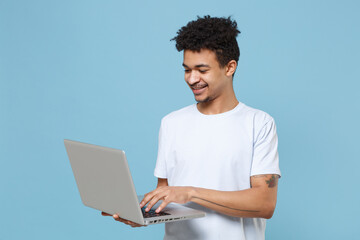 Wall Mural - Smiling young african american guy in casual white t-shirt posing isolated on pastel blue wall background studio portrait. People lifestyle concept. Mock up copy space. Working on laptop pc computer.