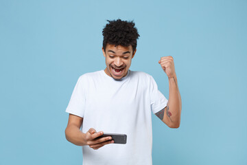 Wall Mural - Happy young african american guy in casual white t-shirt posing isolated on pastel blue wall background. People lifestyle concept. Mock up copy space. Play game with mobile phone doing winner gesture.