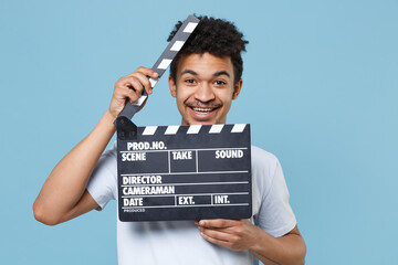 Wall Mural - Smiling young african american guy in casual white t-shirt posing isolated on pastel blue background studio. People lifestyle concept. Mock up copy space. Hold classic black film making clapperboard.