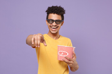 Wall Mural - Laughing african american guy in yellow t-shirt 3d glasses isolated on violet background. People in cinema, lifestyle concept. Watching movie film hold bucket of popcorn, point index finger on camera.