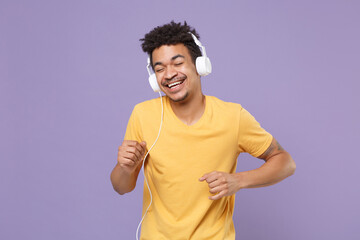 Wall Mural - Cheerful young african american guy in casual yellow t-shirt isolated on violet wall background. People lifestyle concept. Mock up copy space. Listen music with headphones keeping eyes closed dancing.