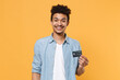 © ViDi Studio - Smiling young african american guy in casual blue shirt posing isolated on yellow wall background studio portrait. People sincere emotions lifestyle concept. Mock up copy space. Hold credit bank card.