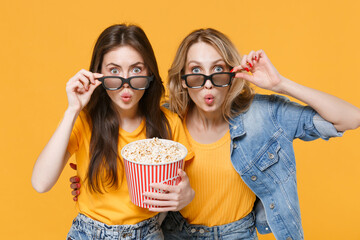 Shocked amazed young women girls friends in denim clothes, 3d glasses isolated on yellow background. People sincere emotions in cinema lifestyle concept. Watching movie film hold bucket of popcorn.