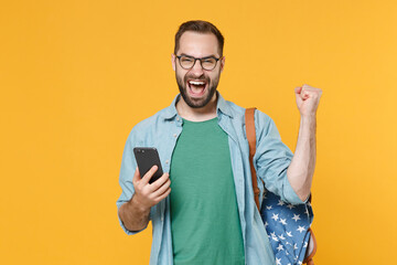 Joyful young man student in casual clothes glasses with backpack posing isolated on yellow background. Education in high school university college concept. Using mobile phone doing winner gesture.