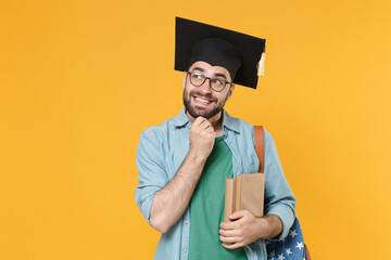 Wall Mural - Pensive young man student in graduation cap glasses with backpack hold books isolated on yellow background. Education in high school university college concept. Put hand prop up on chin, looking up.