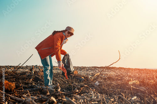 Fotomural A young Caucasian female volunteer in a jacket collects garbage on a dirty beach