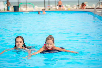 Wall Mural - Two little sisters girls are swimming in a large pool with clear blue water of mine near the hotel on the background of the sea and the beach. Concept vacation tropical hot country with children