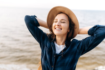 Wall Mural - Close-up portrait of a gorgeous smiling caucasian young woman whose posing in a hat near the sea in a positive mood