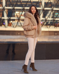 Full length outdoor portrait of young elegant woman wearing casual pullover, trousers and coat with fur hood standing, posing and walking at city street on a sunny winter day