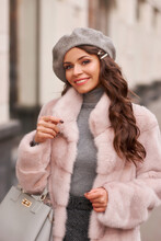 Beautiful Caucasian Woman With Long Brunette Hair In Pink Fur Coat And Gray Beret Walking In City. Outdoor Portrait. Fashionable Luxury Rich Girl