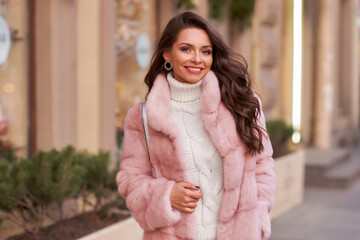 Happy smiling woman outdoor winter portrait. Pretty young rich female in white casual costume and pink fur coat with long wavy hair and makeup walking city street