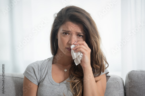 Young beautiful girl sitting at home on the sofa hugging the pillow, watching sad movie on the television or tv and cry wiping away tears with a tissue close up portrait