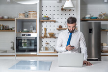 Young Bearded Business Man In Shirt With Neck Tie Standing In The Kitchen Holding Phone And Reading The Statistic On The Laptop Waiting For The Breakfast Before He Go To The Job In The Company