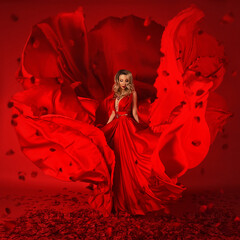A gorgeous blonde girl in a luxurious flying red silk dress with a deep slit standing barefoot on a red background with rose petals.