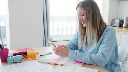 Wall Mural - A pleased young woman wearing glasses is using her smartphone working in the kitchen at home