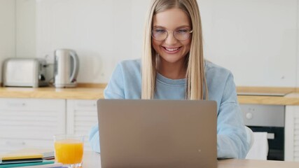 Sticker - A happy satisfied young blonde woman wearing glasses is using her laptop working in the white kitchen at home